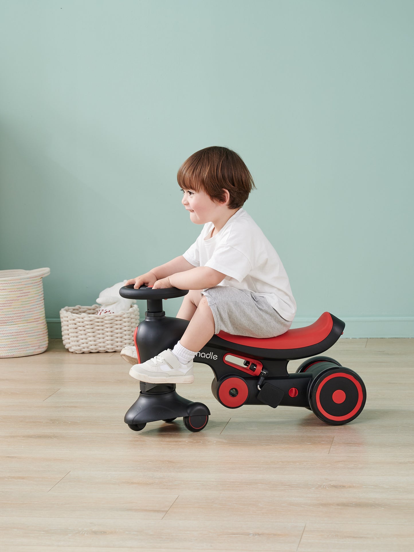 A child happily sits on a black and red ride-on toy indoors, smiling to the side.