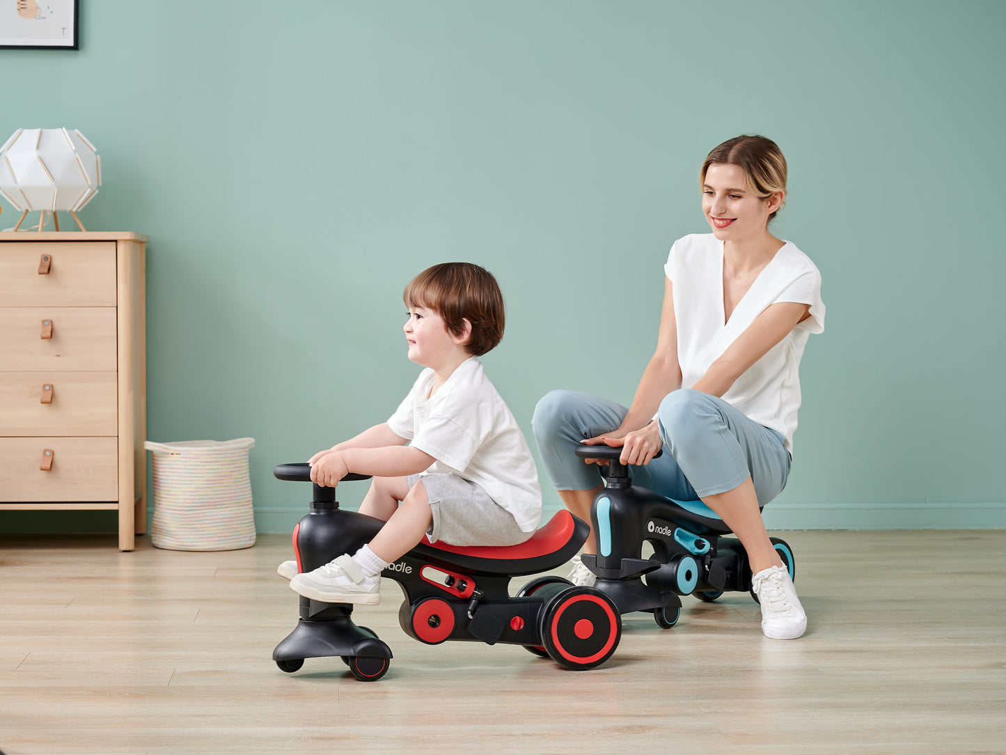 A woman and child sit on colorful ride-on toys, both smiling in a bright indoor setting.