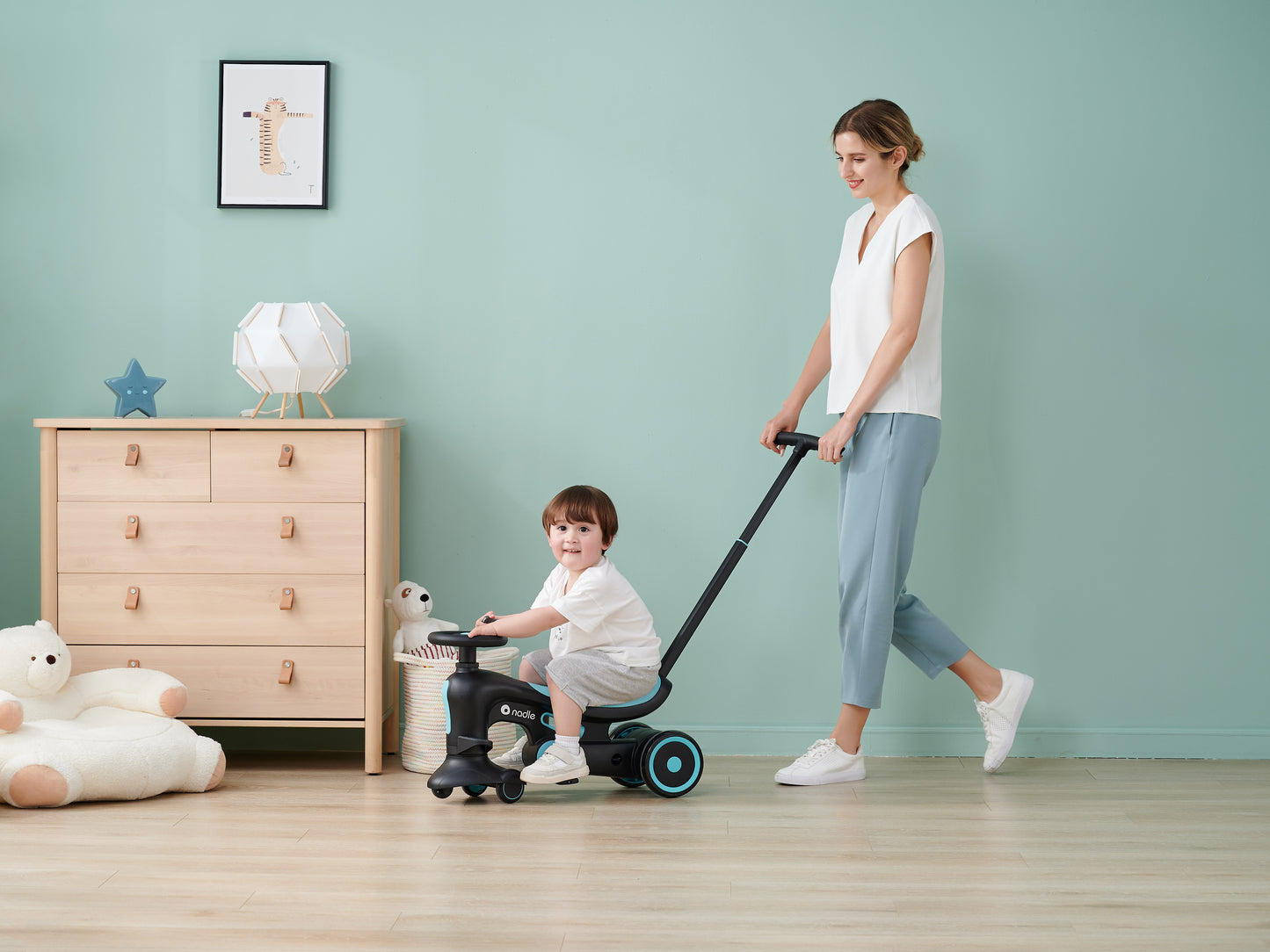 A woman happily pulls a child on a ride-on toy indoors, with a light green wall behind them.