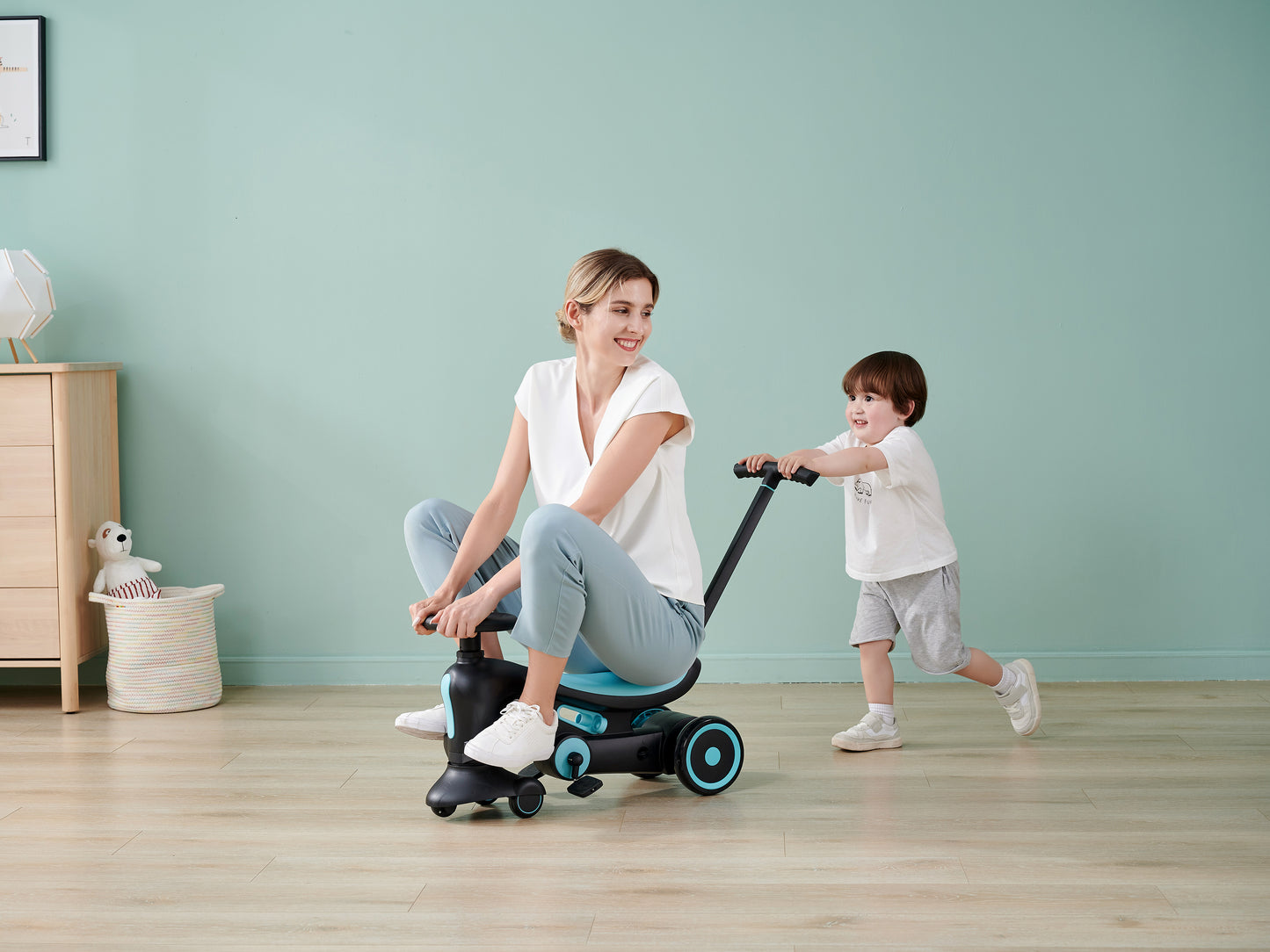 A woman sits on a toy while a child playfully pushes her indoors.
