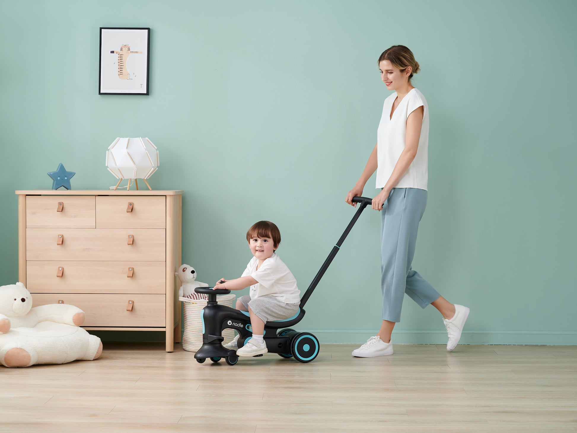 A woman happily pulls a child on a ride-on toy indoors, with a light green wall behind them.