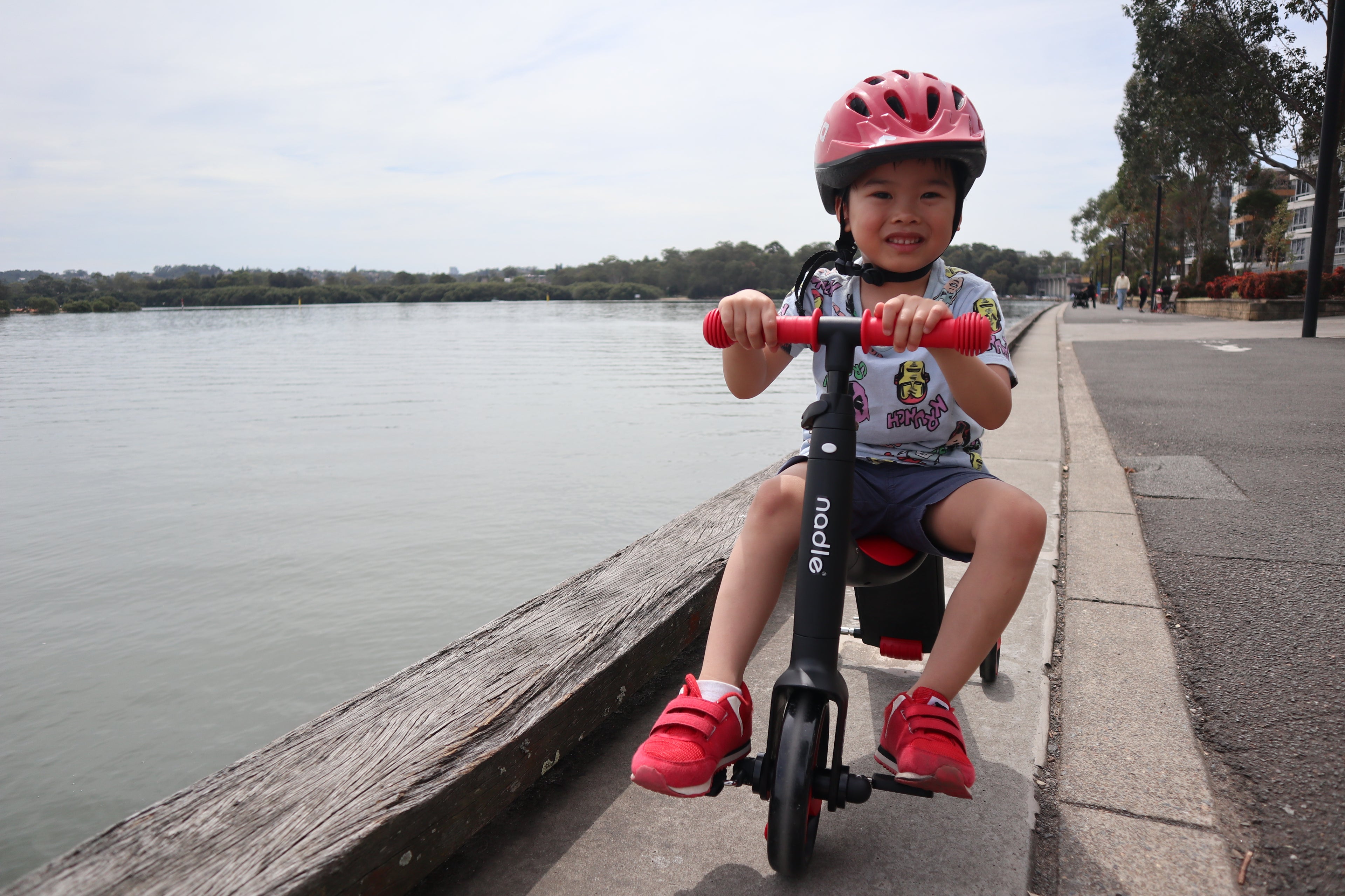 A smiling child with a helmet rides a scooter along a waterfront path.