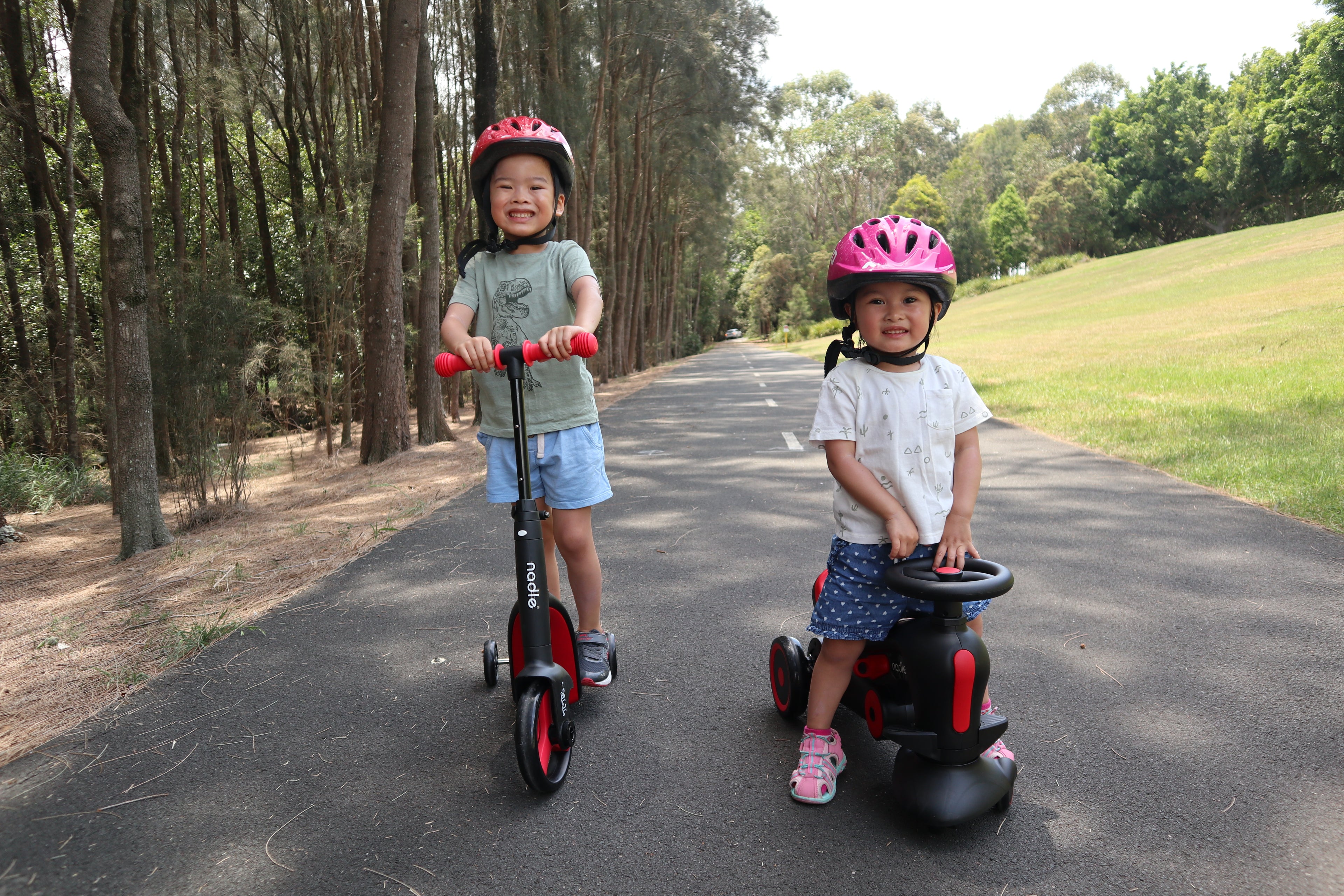 Two smiling children on a scooter and ride-on toy, wearing helmets on a tree-lined path.