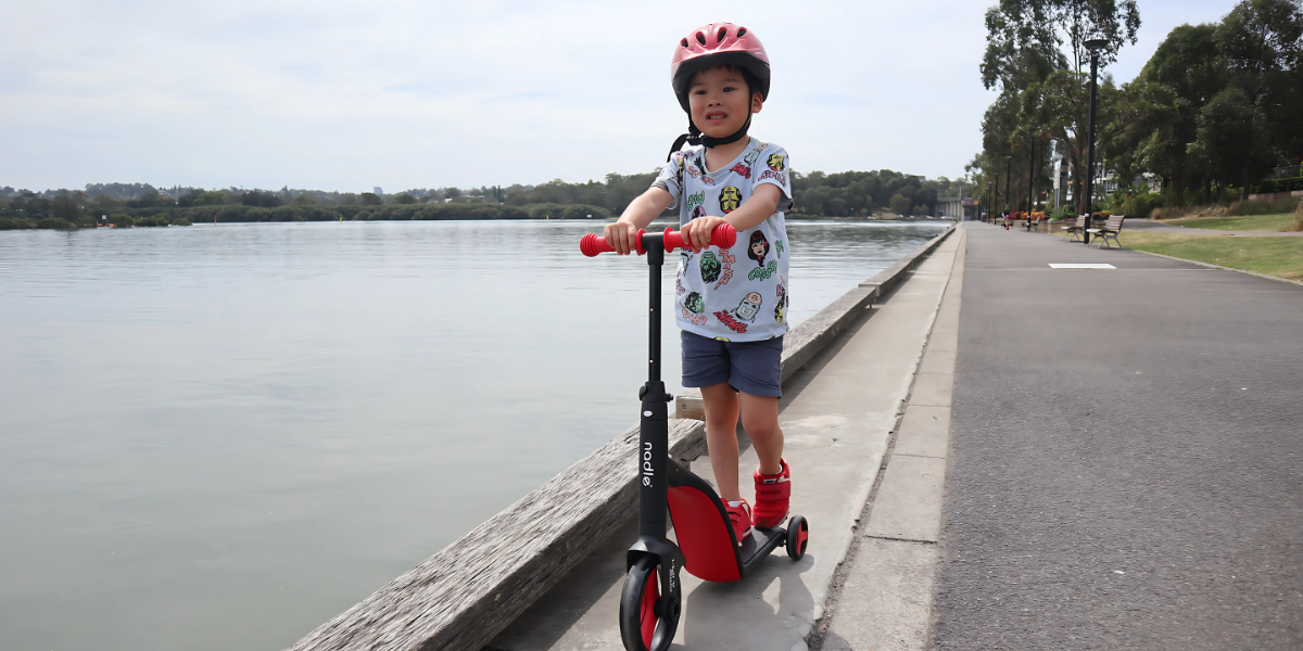 Child on a scooter by the water, wearing a helmet and colorful shirt.